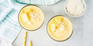 Two glasses of creamy orange smoothie with pineapple chunks and shredded coconut on top, placed on a light gray surface next to a small bowl of shredded coconut, a blue and white striped cloth, and two yellow and white striped straws.