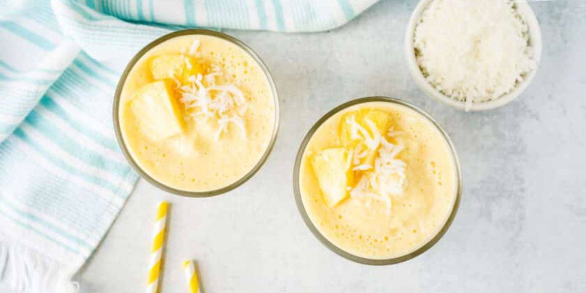Two glasses of creamy orange smoothie with pineapple chunks and shredded coconut on top, placed on a light gray surface next to a small bowl of shredded coconut, a blue and white striped cloth, and two yellow and white striped straws.