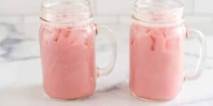 Two mason jars filled with ice and pink-colored liquid, placed on a white marble surface. The jars have handles, and the drink appears to be a cold, creamy beverage.