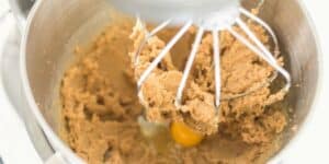 A stand mixer with a wire whisk attachment is shown mixing brown cookie dough. An egg is visible amidst the mixture in the stainless steel mixing bowl. The dough appears partially blended.