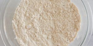 A close-up view of a glass mixing bowl filled with a dry mixture of flour and seasoning. The texture shows small clumps and granules, suggesting the initial stages of a baking or cooking preparation.