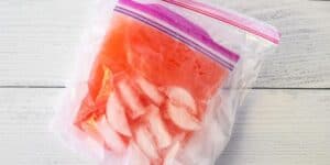 A sealed plastic bag filled with a red liquid and pieces of ice sits on a white wooden surface. The bag has a pink and purple zipper at the top.
