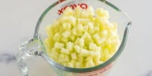 A clear glass measuring cup filled with small, diced pale yellow or green pieces, possibly chopped fruit or vegetables, sits on a white surface. Red measurement markings are visible on the side of the cup.