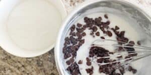 A metal bowl filled with yogurt and chocolate chips being stirred with a whisk is on a granite countertop. Next to it is a smaller white bowl containing plain yogurt.