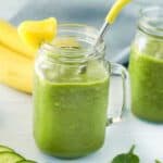 Two mason jars filled with green smoothies, garnished with pineapple pieces and a yellow straw, are on a white surface. In the background, there are bananas, a small bowl of strawberries, and cucumber slices scattered around. A light blue cloth is draped behind.