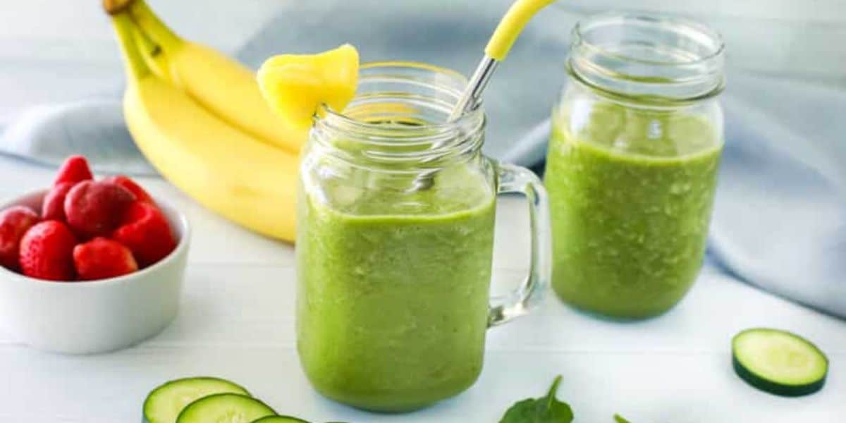 Two mason jars filled with green smoothies, garnished with pineapple pieces and a yellow straw, are on a white surface. In the background, there are bananas, a small bowl of strawberries, and cucumber slices scattered around. A light blue cloth is draped behind.