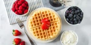 A breakfast setup features a plate with two waffles topped with sliced strawberries. Surrounding it are bowls of raspberries, blueberries, and whipped cream. A small pitcher of syrup and a few whole strawberries are also placed nearby, along with a fork.