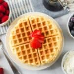 A plate with two golden waffles topped with sliced strawberries. Surrounding the plate are bowls of fresh raspberries, blueberries, and whipped cream. A jar of syrup is also nearby, along with scattered whole strawberries. A fork and a striped cloth napkin are beside the setting.