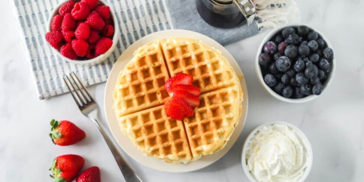 A plate with two golden waffles topped with sliced strawberries. Surrounding the plate are bowls of fresh raspberries, blueberries, and whipped cream. A jar of syrup is also nearby, along with scattered whole strawberries. A fork and a striped cloth napkin are beside the setting.