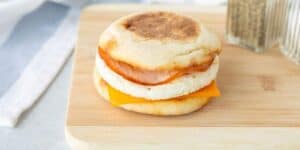 A breakfast sandwich on a light wooden cutting board. The sandwich consists of an English muffin filled with a slice of cheddar cheese, a round egg patty, and a piece of ham. Two shakers, likely for salt and pepper, are visible in the background.