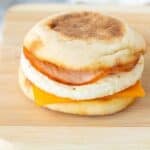A breakfast sandwich on a wooden board featuring an English muffin, egg patty, slice of cheddar cheese, and a piece of ham. Salt and pepper shakers and a striped napkin are in the background.