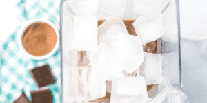 A blender filled with ice cubes sits on a countertop. In the background, there is a blurred checkered cloth, a bowl with brown powder, and pieces of chocolate. The scene suggests preparation for making a Frozen Hot Chocolate Recipe as delightful as Olaf!