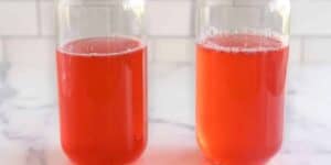 Two clear glass jars, each filled with a bright red liquid, are placed on a white surface. The background features a white tiled wall.