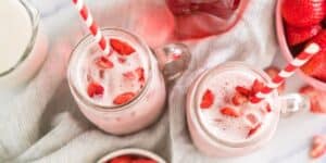 Two mason jars filled with strawberry milk, topped with fresh strawberry pieces and striped red-and-white straws. Nearby are a bowl of whole strawberries, a glass pitcher of milk, and a jar of pink liquid, all set on a soft striped cloth.