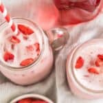 Two mason jars filled with pink strawberry milk, garnished with strawberry slices and red-and-white striped straws. The jars are placed on a white cloth, surrounded by a bowl of fresh strawberries and a glass container of strawberry syrup.