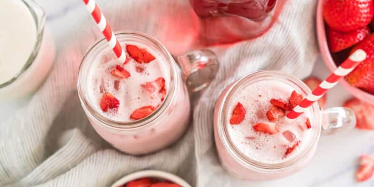 Two mason jars filled with pink strawberry milk, garnished with strawberry slices and red-and-white striped straws. The jars are placed on a white cloth, surrounded by a bowl of fresh strawberries and a glass container of strawberry syrup.