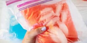 A hand with pale blue nail polish holding a clear plastic zip-lock bag filled with red liquid and ice cubes. The background includes another similar bag containing blue liquid, placed on a light-colored surface.