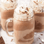 Three glass mugs filled with creamy chocolate milkshakes topped with whipped cream and chocolate shavings. A bowl of cocoa powder, a checkered cloth, and two metal spoons are nearby, creating a scene like a Frozen Hot Chocolate Recipe as Delightful as Olaf! In the background, a chocolate bar and scattered ice cubes add charm.