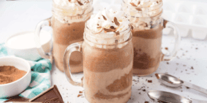 Three glass mugs filled with creamy chocolate milkshakes topped with whipped cream and chocolate shavings. A bowl of cocoa powder, a checkered cloth, and two metal spoons are nearby, creating a scene like a Frozen Hot Chocolate Recipe as Delightful as Olaf! In the background, a chocolate bar and scattered ice cubes add charm.