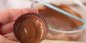 A hand is holding a chocolate cup filled with smooth melted chocolate. In the background, there is a glass bowl containing more melted chocolate with a light blue spatula resting inside it. The setting appears to be a kitchen or baking area.