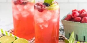 Two glasses of berry lemonade are garnished with fresh berries, lime slices, and mint leaves on a checkered cloth. A basket of raspberries, sliced limes, and striped straws are placed nearby. The refreshing drinks are chilled with ice and have a vibrant pink color.