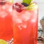 Two tall glasses filled with a pink beverage, ice, and garnished with lime slices, blackberries, and mint leaves sit on a checked cloth. A box of fresh raspberries, sliced limes, striped straws, and mint leaves are arranged around the glasses.