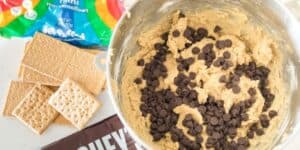 A mixing bowl filled with cookie dough and chocolate chips sits on a white countertop. Nearby are ingredients including a partially opened bag of mini marshmallows, graham crackers, and a bar of Hershey's chocolate.