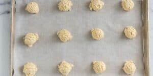 A baking tray lined with parchment paper holds 12 evenly spaced, round scoops of cookie dough, arranged in a 4x3 grid. The tray is placed on a marble surface.