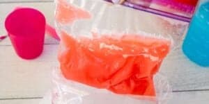 A plastic zipper bag filled with red liquid is being poured into another plastic bag. A pink cup sits on the left, and a blue liquid container is partially visible on the right. The background has a white wooden surface.