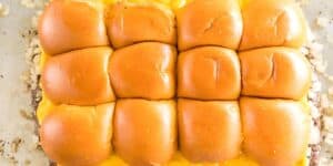 A tray of freshly baked cheeseburger sliders, topped with shiny golden-brown buns. The sliders are neatly arranged in a 3x4 grid, with melted cheese visible between the buns. The buns appear fluffy, and there are small bits of finely chopped onions around them.