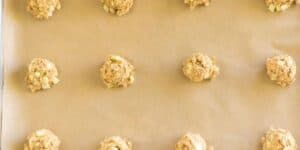 Twelve evenly spaced, unbaked cookie dough balls with visible chunks of ingredients are placed on a sheet of parchment paper. The dough balls are arranged in a grid pattern on the baking tray, ready to be baked.