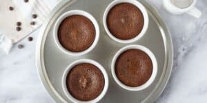 A round baking tray with four chocolate soufflés in white ramekins. The tray is placed on a gray marble countertop. Nearby are a white cloth with brown stripes, a few chocolate chips, and a white sifter.