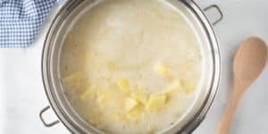 A stainless steel pot filled with a creamy, cheesy potato soup that rivals Le Cellier’s iconic dish sits on a white surface. A blue and white checkered cloth is partially visible in the background, and a wooden spoon rests beside the pot.