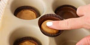 A hand with white-painted nails pressing down on a peanut butter cup in a cupcake liner inside a muffin tray. Other peanut butter cups in liners are visible in the tray.