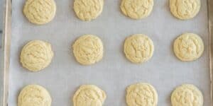 A baking tray with twelve evenly spaced, round cookies on parchment paper. The cookies appear soft and slightly golden with a lightly textured surface.