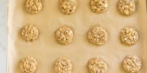 A baking sheet lined with parchment paper holds twelve evenly spaced oatmeal cookies. The cookies are golden brown and contain small chunks of what appears to be apple, giving them a rustic, homemade appearance.