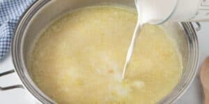 A metal pot is filled with a creamy yellow mixture, likely cheesy potato soup that rivals Le Cellier’s iconic dish, as a measuring cup pours cream or milk into it. A blue and white checkered cloth is partially visible in the background.