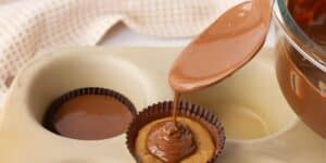 A spoon pours melted chocolate over a peanut butter cup that sits in a muffin tin. Another partially filled peanut butter cup is visible in the background, with a cloth in a checkered pattern behind the tin.