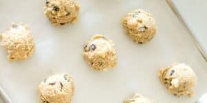 Six scoops of chocolate chip cookie dough sit evenly spaced on a parchment paper-lined baking sheet, ready to be baked. The dough is studded with visible chunks of chocolate.