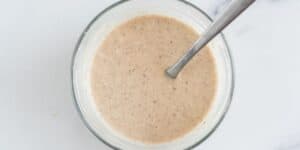 A small clear glass bowl filled with a beige, speckled mixture. A metal spoon is resting inside the bowl, and the bowl is placed on a white surface.