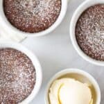 Three small white ramekins filled with chocolate lava cake are dusted with powdered sugar and placed on a white surface. A bowl of vanilla ice cream is on the side, along with an extra scoop of ice cream, a gold spoon, and a few scattered chocolate chips.