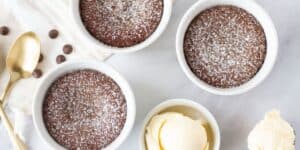 Three small white ramekins filled with chocolate lava cake are dusted with powdered sugar and placed on a white surface. A bowl of vanilla ice cream is on the side, along with an extra scoop of ice cream, a gold spoon, and a few scattered chocolate chips.