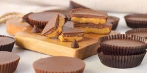 Assorted chocolate peanut butter cups are displayed on a wooden board and surrounding surface. Some cups are cut in halves, revealing the peanut butter filling. Two chocolate kisses are also present among the treats.