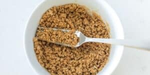 A white bowl filled with crumbly brown mixture. A fork is placed in the bowl, partially mixing the contents. The mixture appears to be coarse and textured, likely a streusel or crumb topping used for baking. The background is a white surface.