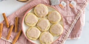 A plate of seven frosted cookies is placed on a pink napkin. Next to the plate are some cinnamon sticks, two tea bags labeled "Chai Tea," and a cup partially visible in the upper left corner. The cookies appear to have a light brown glaze on top.