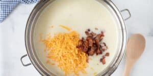 A stainless steel pot contains a creamy cheesy potato soup that rivals Le Cellier’s iconic dish. Shredded cheddar cheese and crumbled bacon are visible on the surface. A wooden spoon rests beside the pot on a white marble countertop, with a blue and white checked cloth partially visible.