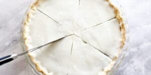 A top-down view of a cream pie being sliced. The pie is in a clear glass dish with a smooth, white topping. One slice is being cut with a knife, and the pie is divided into equal portions. The background is a pale marble surface.