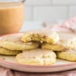 A plate of frosted sugar cookies are arranged on a pink-rimmed plate, with one cookie partially eaten atop the stack. In the background, a glass mug of coffee sits, surrounded by cinnamon sticks and a pink cloth napkin on a white kitchen counter.