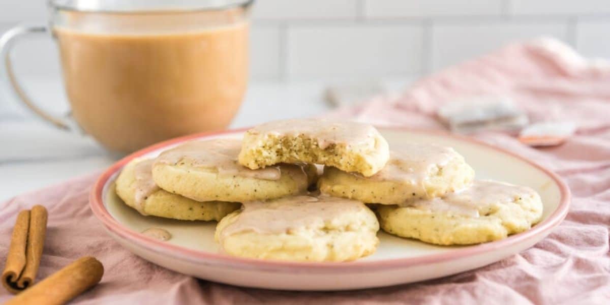 A plate of frosted sugar cookies are arranged on a pink-rimmed plate, with one cookie partially eaten atop the stack. In the background, a glass mug of coffee sits, surrounded by cinnamon sticks and a pink cloth napkin on a white kitchen counter.