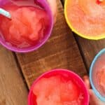 A top-view image of four colorful slushies in round cups on a wooden surface. The slushies are various shades of red and pink, and each cup has a striped straw in it. The cups are purple, yellow, blue, and pink. A hand is holding the blue cup at the bottom.